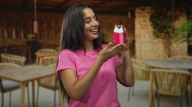 Gülümseyen kadın kameraya bakarken güneşli bir açık hava restoranının terasında iki eliyle pembe şişe sunar..