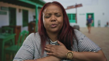 Yeşil binanın girişinde göğsünü sıkıca tutan kadın çizgili gömlek ve tahta kol saati takıyor; kalp krizi acısı.