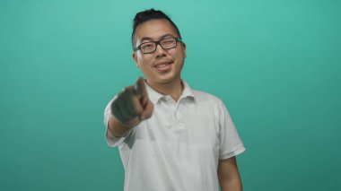 Genç adam stüdyoda kamerayı işaret ediyor, tepe örtüsü kapalı gülümsüyor, beyaz polo tişört giyiyor; dostça bir davet..