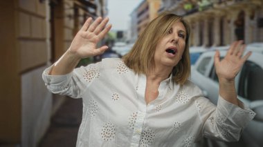 Orta yaşlı beyaz kadın, park halindeki arabaları olan kalabalık bir şehir sokağında ellerini kaldırarak gözlerini koruyor..