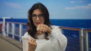 Parlak bir yolcu güvertesinde, okyanus arkaplanlı bir korkuluğun önünde yumruğunu yumruklayan kadın, kararlılık..