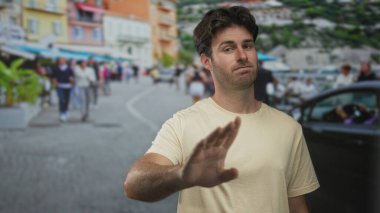 Göğüs göğüse ve avuç içi havaya kaldırılmış bir adam kalabalık bir şehir caddesinde kafeler ve park edilmiş arabalarla, bej tişört giymiş, şüphecilik.