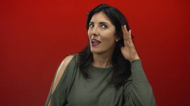 Sağ kulağını kavrayan ve stüdyoda öne doğru eğilen parlak kırmızı arka planlı bir kadın açık bir şekilde belli belirsiz bir ses duymak için; merak.