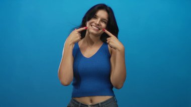 Stüdyoda gülümseyen yanakları gösteren mavi arka planı ve görünür göbeği olan bir kadın..