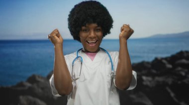 Beyaz önlük ve mavi steteskop giyen genç Afrikalı Amerikalı kadın doktor stüdyoda yumruklarını kaldırdı..