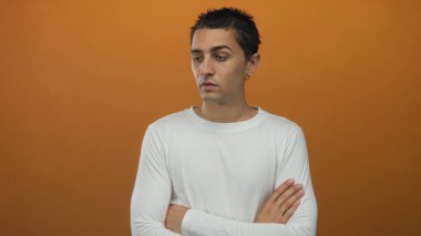 İzole edilmiş turuncu arka planda kollarını kavuşturmuş genç bir adam beyaz gömlek giyerken düşünceli görünüyor..