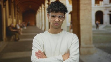 Eski kasaba caddesinde kolları çapraz olan genç İspanyol adam dar kemerli sütun geçidi altında; kafa karışıklığı şüphesi belirsizlik.