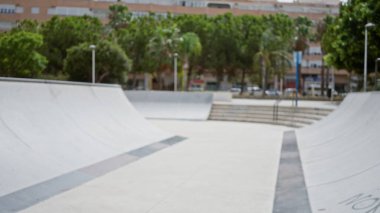 Bulanık ağaçlar ve binalarla dolu bir açık hava parkında beton rampalar ve raylar ile paten parkı çözünmüş ön plan; arka plan telif alanı sakinliği.