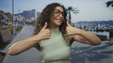 Kıvırcık saçlı ve gözlüklü kadın güneşli sahil caddesinde çifte onay veriyor. İyimserlik teşvik..