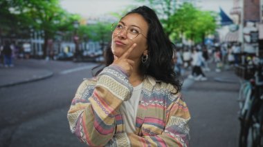 Genç kadın işaret parmağı yukarı doğru işaret ediyor, temiz gözlük ve halka küpelerle gülümsüyor, desenli ceket ve beyaz tişört giyiyor; neşeli bir merak.