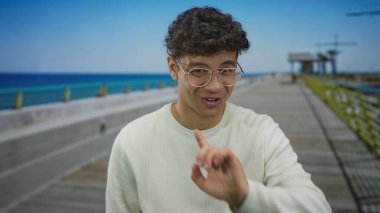 Gözlüklü bir adam beyaz bir süveter giyer manzaralı bir caddede parmağını gösterir; neşeli bir heyecan.