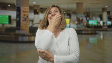 Uzun kollu beyaz gömlek giyen kadın yoğun havaalanı terminalinde ağzını kapatıyor; yorgunluk.