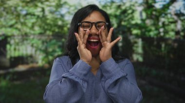 Gözlüklü ve mavi bluzlu kadın, parkta güneşli yeşil yapraklar arasında el kaldırıyor ve gülüyor; saf neşe.