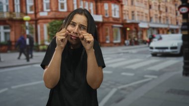 Genç İspanyol kadın gözlerini ovuşturuyor ve yanaklarını bir şehir caddesi yaya geçidinde çekiyor, dışarıda duruyor; üzüntü yorgunluğu.