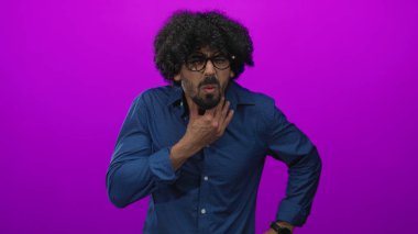 Kıvırcık saçlı ve yuvarlak gözlüklü bir adam stüdyoda boğazını sıkan morumsu bir perde arkasında, eli ağrılı bir boğaz ağrısı olan bir adam..