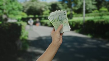Güneşli bir caddede elinde Singapur doları tutan adam şehirde para ve açık hava aktivitelerini ön plana çıkarıyor..