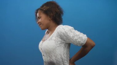 Mavi stüdyoda sırt ağrısı çeken, beyaz örgü hırka giyen ve yan profilde yüzünü buruşturan sınırlı hareketli, ağrılı bir kadın..