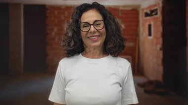 İnşaat alanında güvenle gülümseyen bir kadın. Beyaz tişörtünün içinde dayanıklılık ve güç gösterisi yapıyor. Arka planda tuğla duvarlar var..