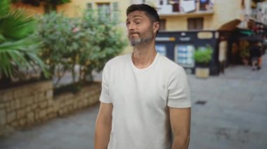 Elleri alnında, gözleri kapalı dar bir sokakta beyaz tişört giyen bir adam..