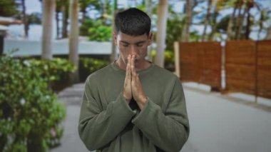 Palmiye ağaçlarıyla kaplı yeşil park yolunda ellerini dudaklarına yapıştıran İspanyol genç adam, huzur yansıması.