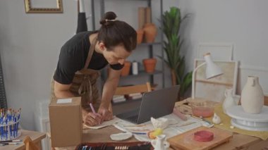 Önlüklü bir adam dizüstü bilgisayarının yanında kalemle yazı masasına yaslanıyor ve atölyedeki çalışma masasında kilden vazolar bulunuyor..