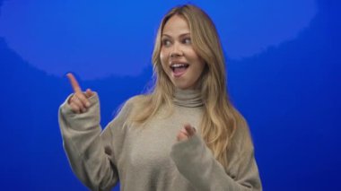 Mavi stüdyoda, neşeli ifadeleri ve görünür elleri olan ve heyecan enerjisi olan bir kadın işaret ediyor..
