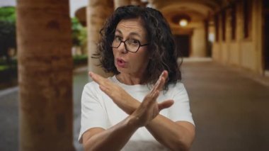 Orta yaşlı kıvırcık saçlı ve gözlüklü İspanyol kadın, eski kasaba sokaklarında reddedilme jesti olarak kollarını çaprazlıyor..