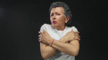 Yaşlı bir kadın, gri saçlı, siyah bir arka planda soğuk görünüyor, kollarını tutuyor ve beyaz bir gömlek giyiyor, soğuk bir rahatsızlık veriyor..