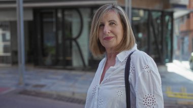 Beyaz bluzlu ve omuz çantalı bir kadın binanın önünde duruyor..