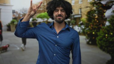 Kaya boynuzları yapan kıvırcık saçlı bir adam el hareketi yapıyor ve süslenmiş yeşil ağaçları olan bir caddede gülümsüyor; neşe dolu bir kutlama enerjisi.