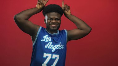 Mavi basketbol formalı adam stüdyoda parmaklarını başının üstüne kaldırıyor ve Los Angeles 'ı 77 numaralı kırmızı duvara yaslıyor..