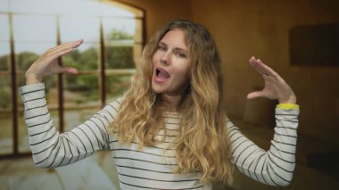 Kadın, kapalı bir sahnede dedikodu yaparken ellerini kaldırıyor. Sarışın, genç bir kadın, pencerenin yanında çizgili bir bluz giyiyor. Ağız hareketi yapıyor..