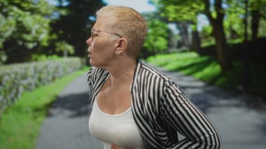 Beyaz kolsuz tişörtlü orta yaşlı bir kadın ve çizgili ceketli. Sırtını, yeşil orman yolunda kalçaları olan bir kadın. Ağrıya dayanıklılık..