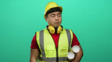 Asyalı genç bir adam inşaat malzemesi giyiyor yeşil arka planla ilgili planlar tutuyor mesleğini gösteriyor ve açık havada mimarlık işine odaklanıyor.