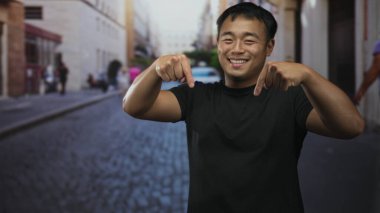 Genç Çinli adam güpegündüz, işlek bir kaldırım taşı caddesinde parmaklarını aşağı doğru işaret ediyor. Mutluluk..