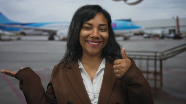Woman showing thumbs up and holding open palm toward airplane at airport terminal outdoors; approval.