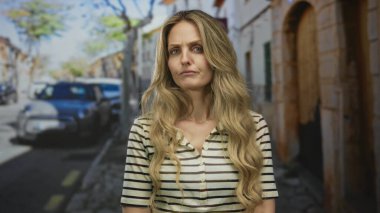 Sokakta duran uzun sarı saçlı, çizgili tişörtlü, arabalarla ve binalarla çevrili bir kadın, şehir ortamında ciddi bir tavır sergiliyor..