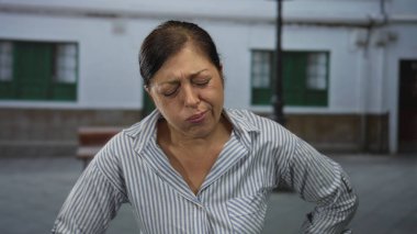 Orta yaşlı İspanyol kadın sırtı aşağıda ve öne doğru eğiliyor. Şehir caddesinde buruşuk yüzüyle; sırt ağrısı.