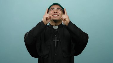 Rahip gibi giyinmiş mutlu bir adam gülümsüyor ve mavi bir arka plana işaret ediyor.