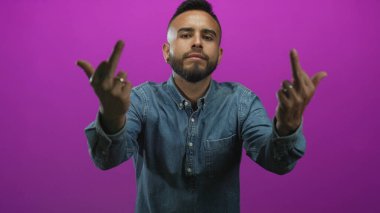 Her iki orta parmağını da kameraya doğru gösteren adam, mor stüdyoda yüzükler ve kot gömlekli, meydan okuyan tavır..