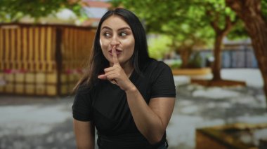 Caddede sessiz kalmak için parmaklarını dudaklarına bastıran kadın ahşap kulübenin ve gölgeli ağaçların yanında, hafif bir gülümsemeyle yan bakıyor; şakacı gizlilik.