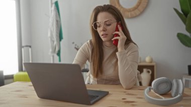 Binadaki ahşap bir masada dizüstü bilgisayara bakarken kırmızı akıllı telefonu kulağına tutan kadın. Konsantrasyon..