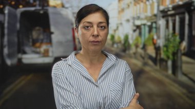 Çizgili tişörtlü bir kadın güneş ışığıyla çevrili şehir sokağında duruyor. Park etmiş minibüs ve mağazalarla dolu. İnkar..