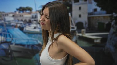 Kadın yan profilde, kayığın yanında beyaz bir kolsuz bluzla taş bir binaya karşı somurtuyor..