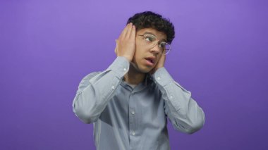 Mavi tişörtlü genç İspanyol adam mor stüdyoda kulaklarını kapatıyor ve yukarı bakıyor..