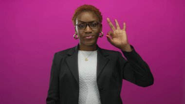 Gözlük ve halka küpe takan bir kadın, parlak pembe bir stüdyo geçmişiyle el işareti yapıyor. Onaylandı..
