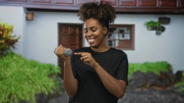 Gülümseyen kadın, yeşil bitkiler ve ahşap kapılı bir binanın girişinde avuç içini gösterirken sağ elinde 1kg dambıl tutuyor..