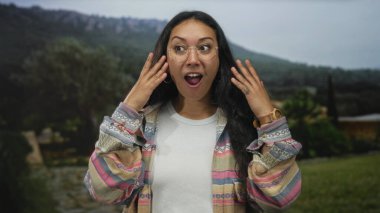 Elden yüze ve ağzı açık bir kadın, gözlük takıyor ve desenli ceket giyiyor, yeşil alan görünen bir ormanda duruyor; sürpriz keşif.