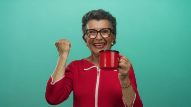 Kırmızı elbiseli, gri saçlı bir kadın elinde kırmızı bir kahve fincanı tutarken gülümsüyor, neşe ve neşe saçıyor..