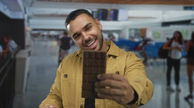 Bir elinde çikolata tutan ve havaalanı terminalinde gülümserken onu inceleyen adam, cezbedici neşe..